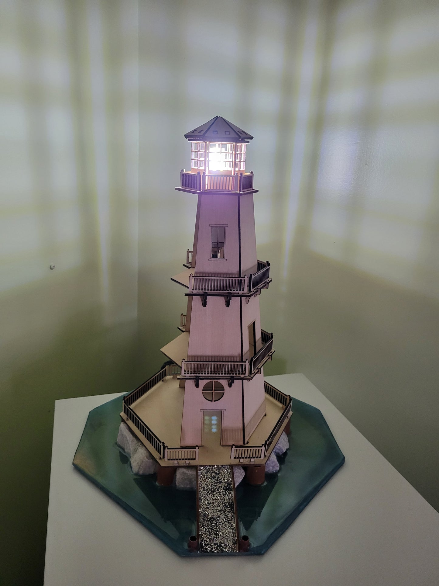Wooden lighthouse model with illuminated lantern room and water base.