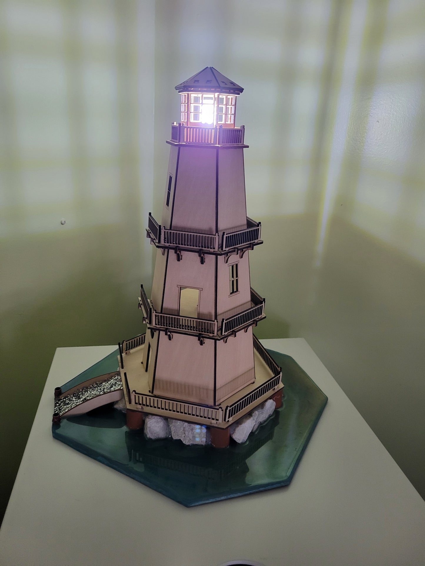 Wooden lighthouse model with illuminated top on a green hexagonal base.