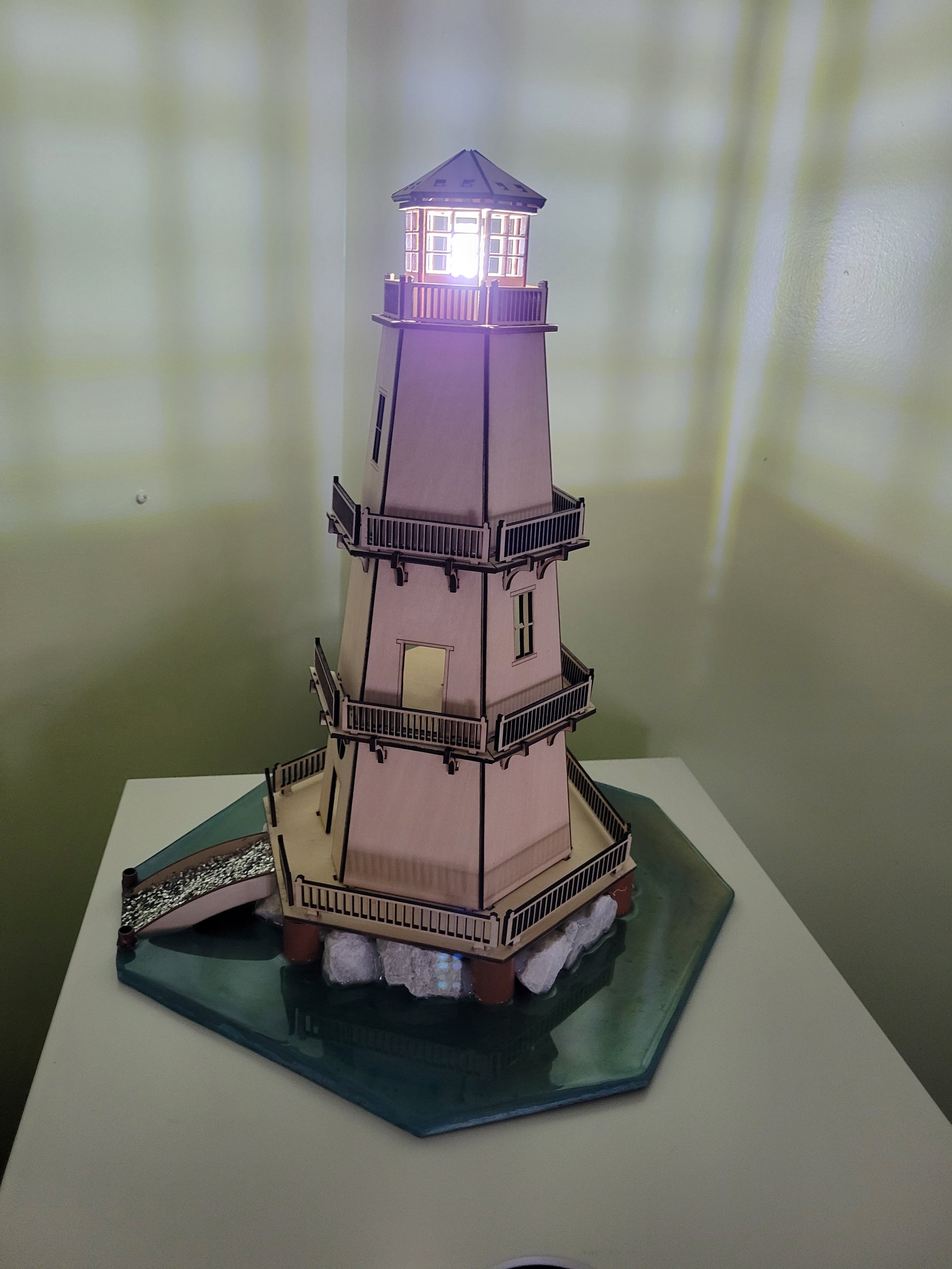Wooden lighthouse model with illuminated top on a green hexagonal base.