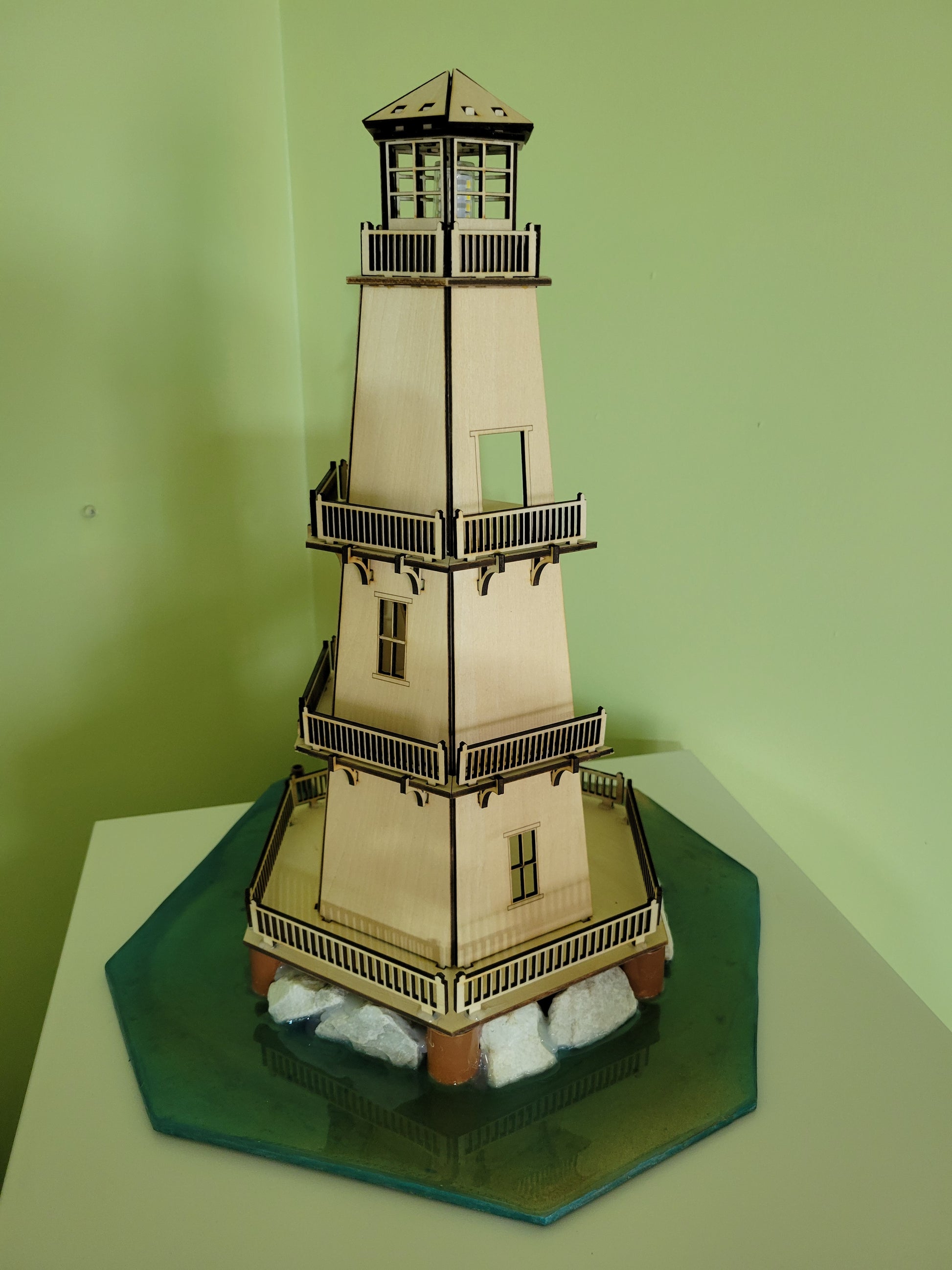 Wooden model lighthouse with stairs, windows, and rocks on a green base.