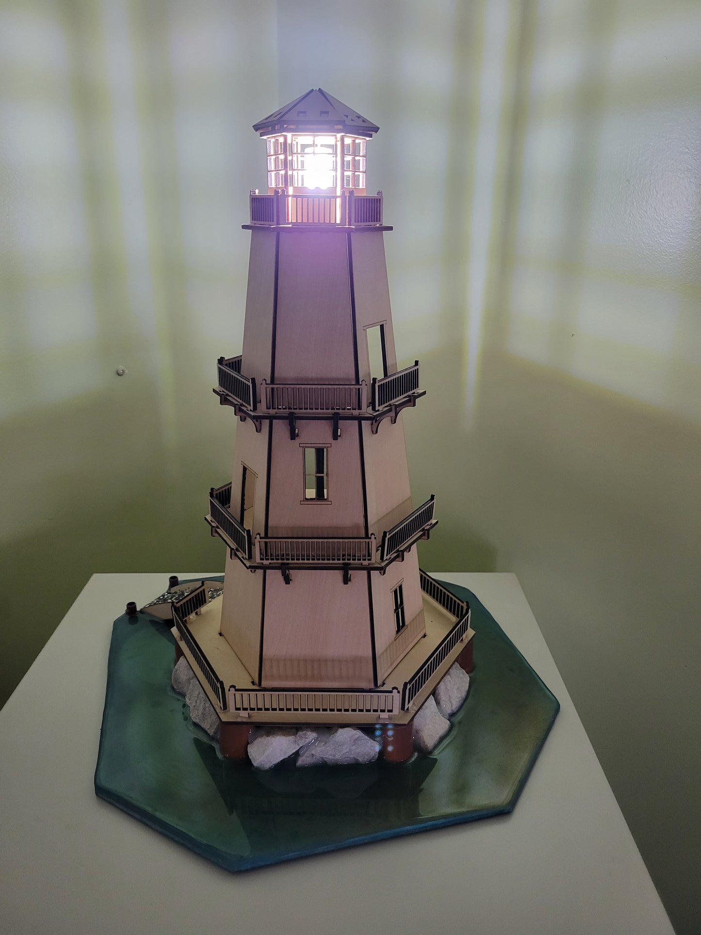 Lighthouse model with illuminated top on a blue-green base with rocks.
