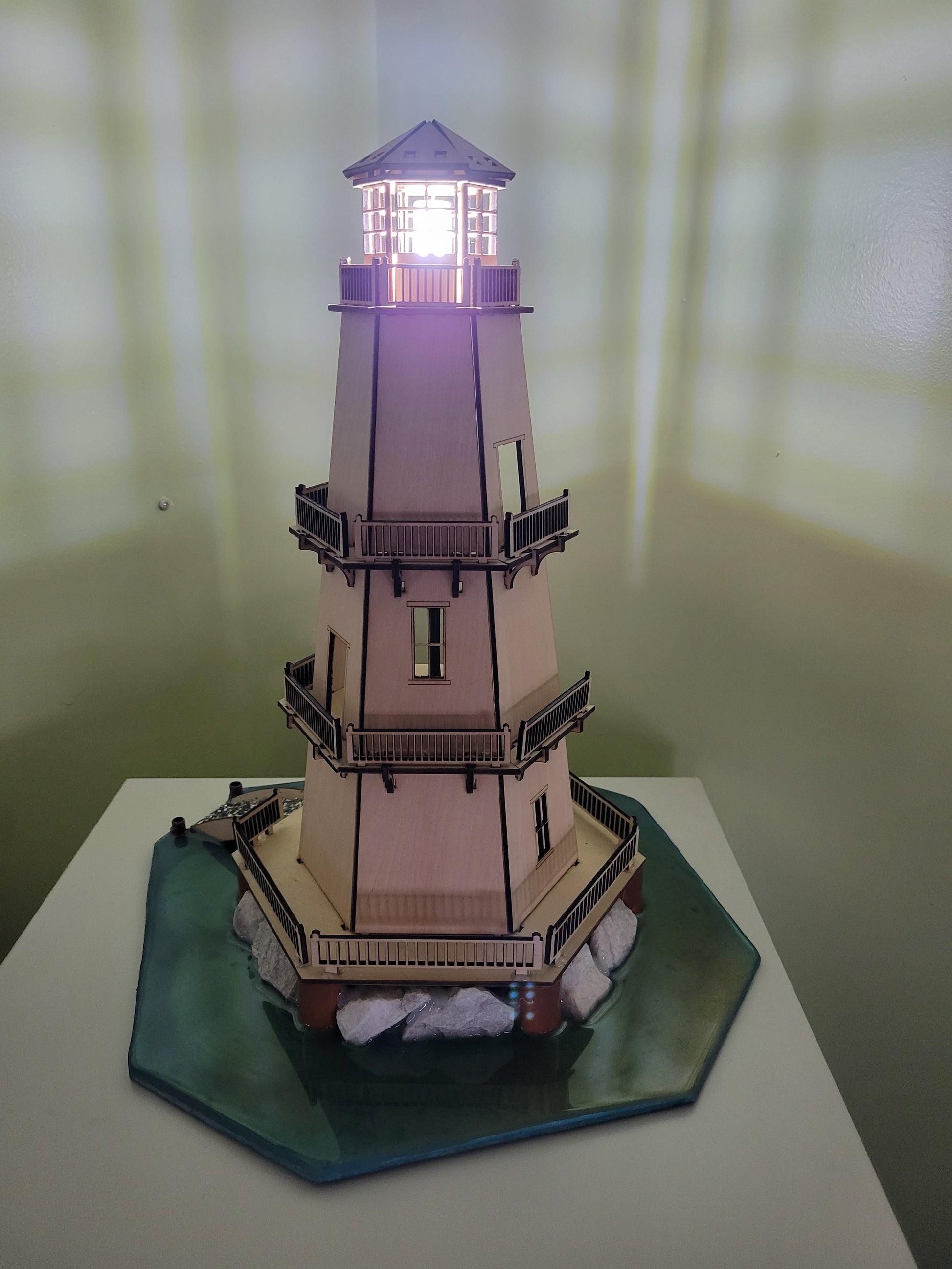 Lighthouse model with illuminated top on a blue-green base with rocks.