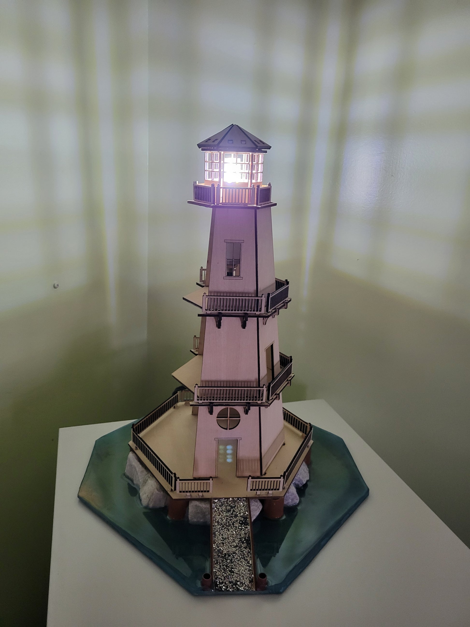 Wooden lighthouse model with illuminated lantern room and water base.