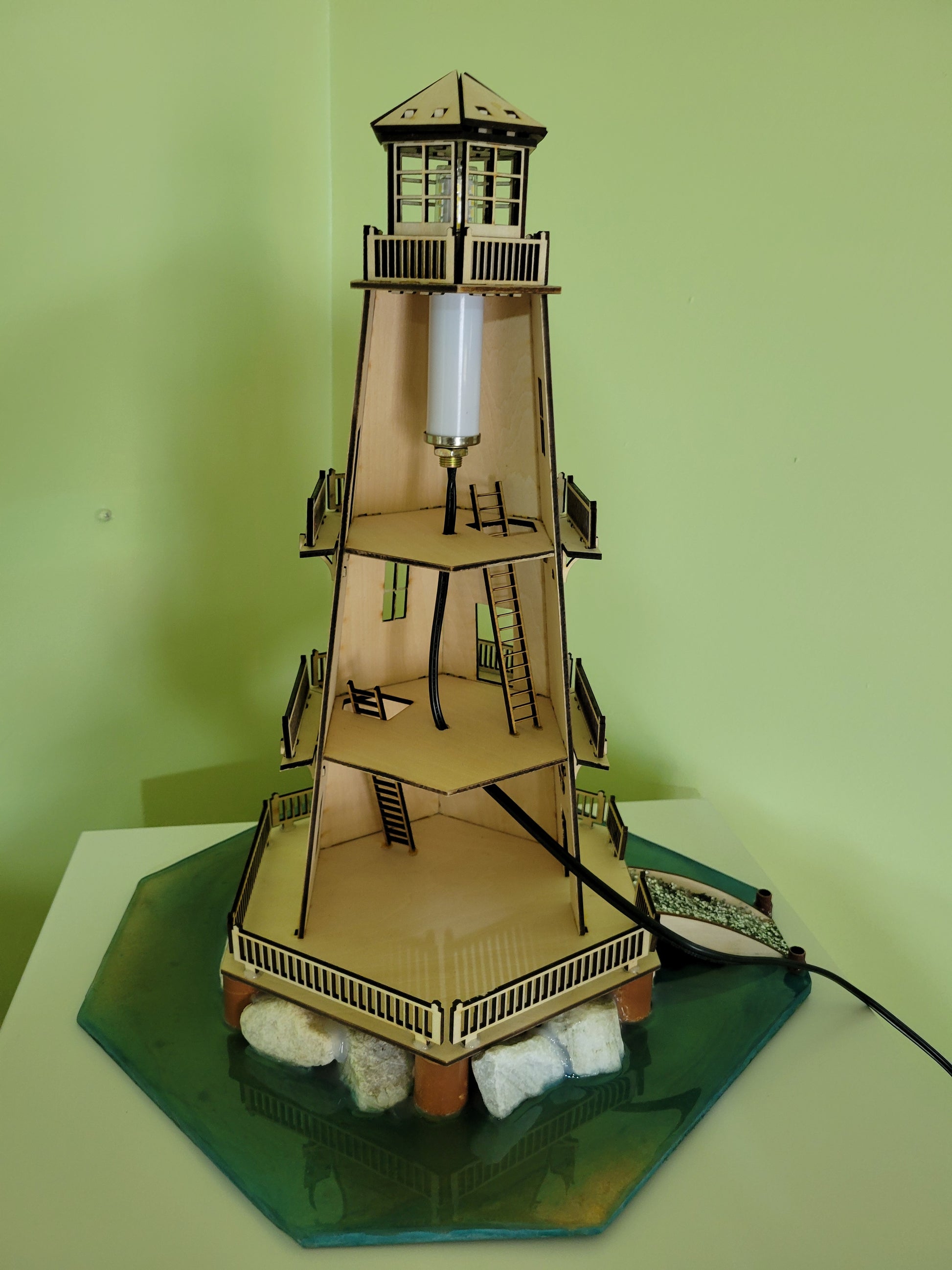Wooden lighthouse model with interior details and faux rocks on a green base.